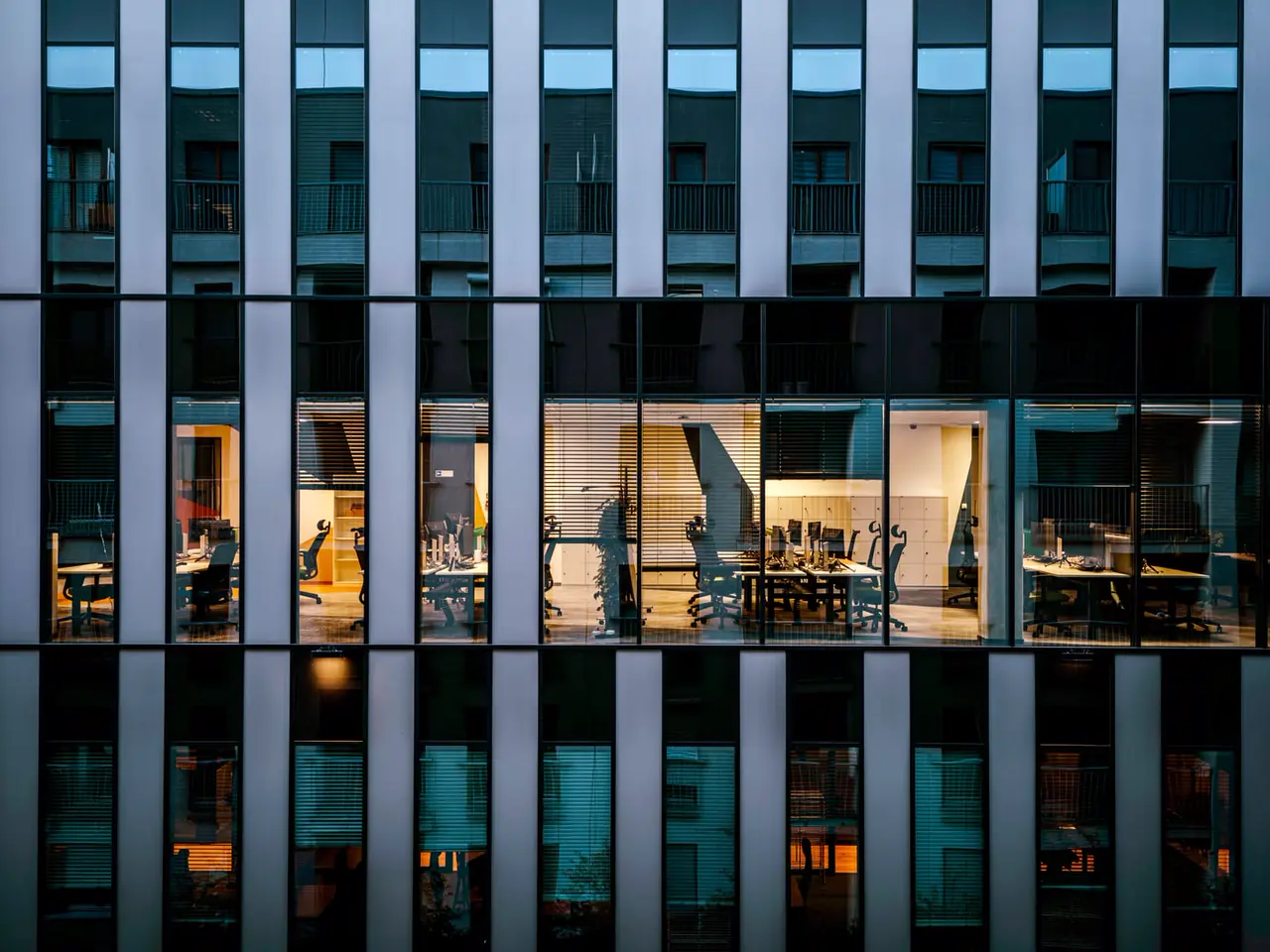 An office floor at dusk, seen through the glass facade, desks and chairs lit and empty.
